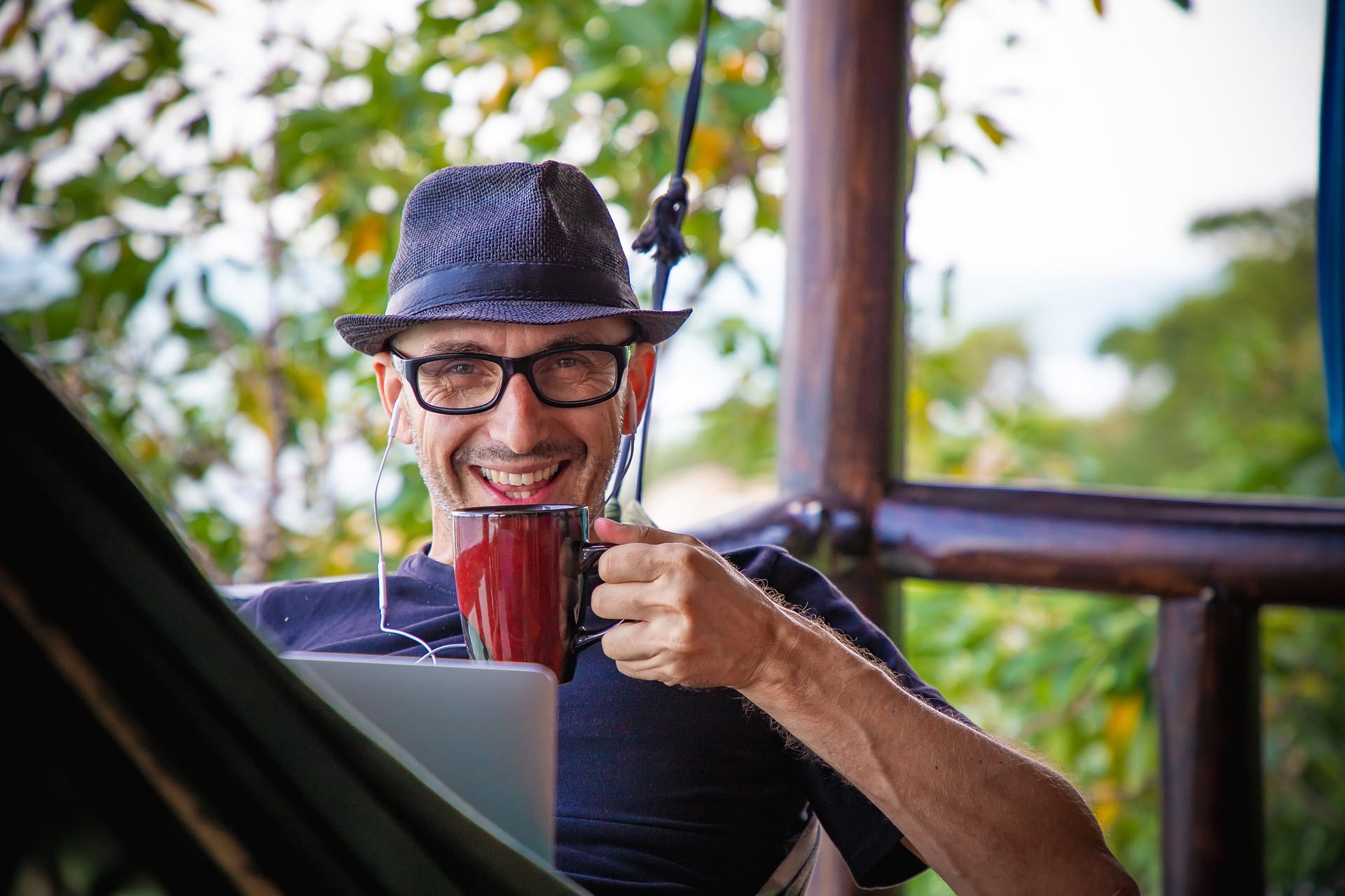  A man wearing a hat and glasses is smiling while holding a cup of coffee and looking at his laptop. The image represents the search query 'Tips to boost charisma without wearing gold amulets'.